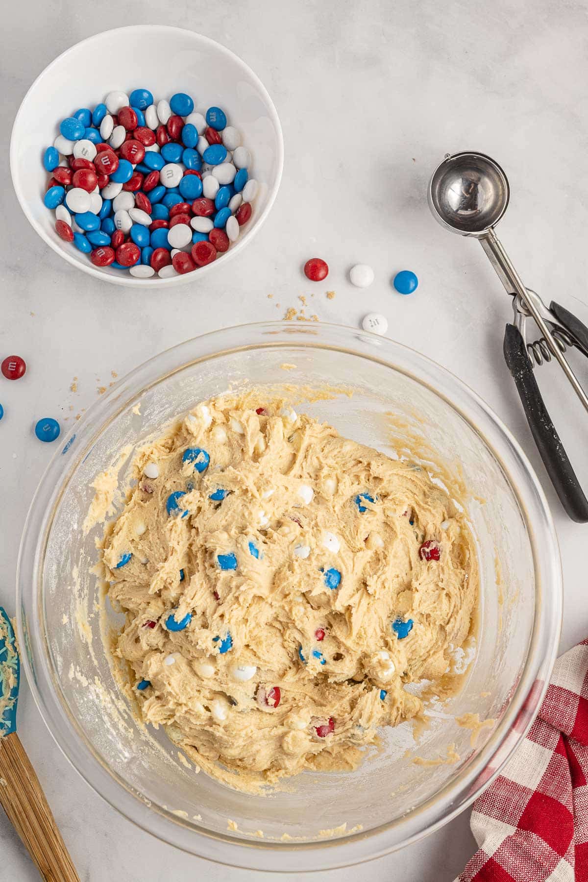 A clear bowl contains cookie dough mixed with red, white, and blue candy-coated chocolates. Nearby are a metal scoop, a white bowl with more chocolates, and a red and white checkered cloth.