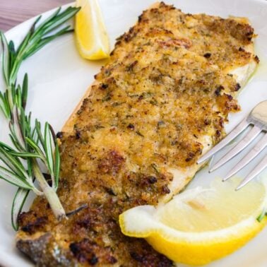 A breaded and seasoned baked fish fillet garnished with a lemon wedge and sprigs of rosemary on a white plate with a fork.