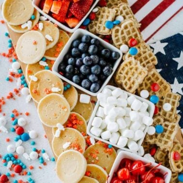 A festive platter with mini pancakes, blueberries, marshmallows, strawberries, small waffles, and red, white, and blue candies, arranged on a wooden board with an American flag backdrop.