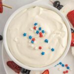 A bowl of sweet cream cheese dip topped with red, white, and blue star-shaped sprinkles. Surrounding the bowl are strawberries, blueberries, and pieces of cake.