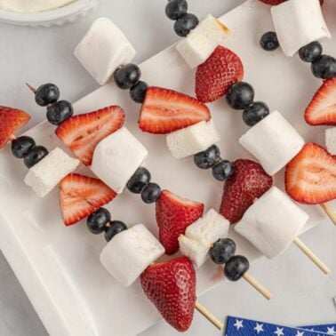 Skewers with strawberries, blueberries, marshmallows, and small cake squares neatly arranged on a white plate.