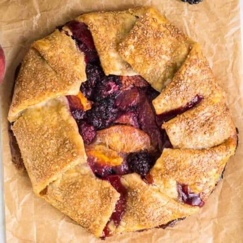 A rustic peach galette featuring a golden, flaky crust filled with a mix of baked berries and peach slices on parchment paper.