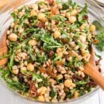 A glass bowl filled with a mixed salad containing chickpeas, arugula, farro, raisins, and sun-dried tomatoes, being tossed with wooden utensils.