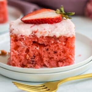 A slice of strawberry jello cake topped with a thin layer of pink frosting and a strawberry slice on a white plate, with a gold fork beside it.