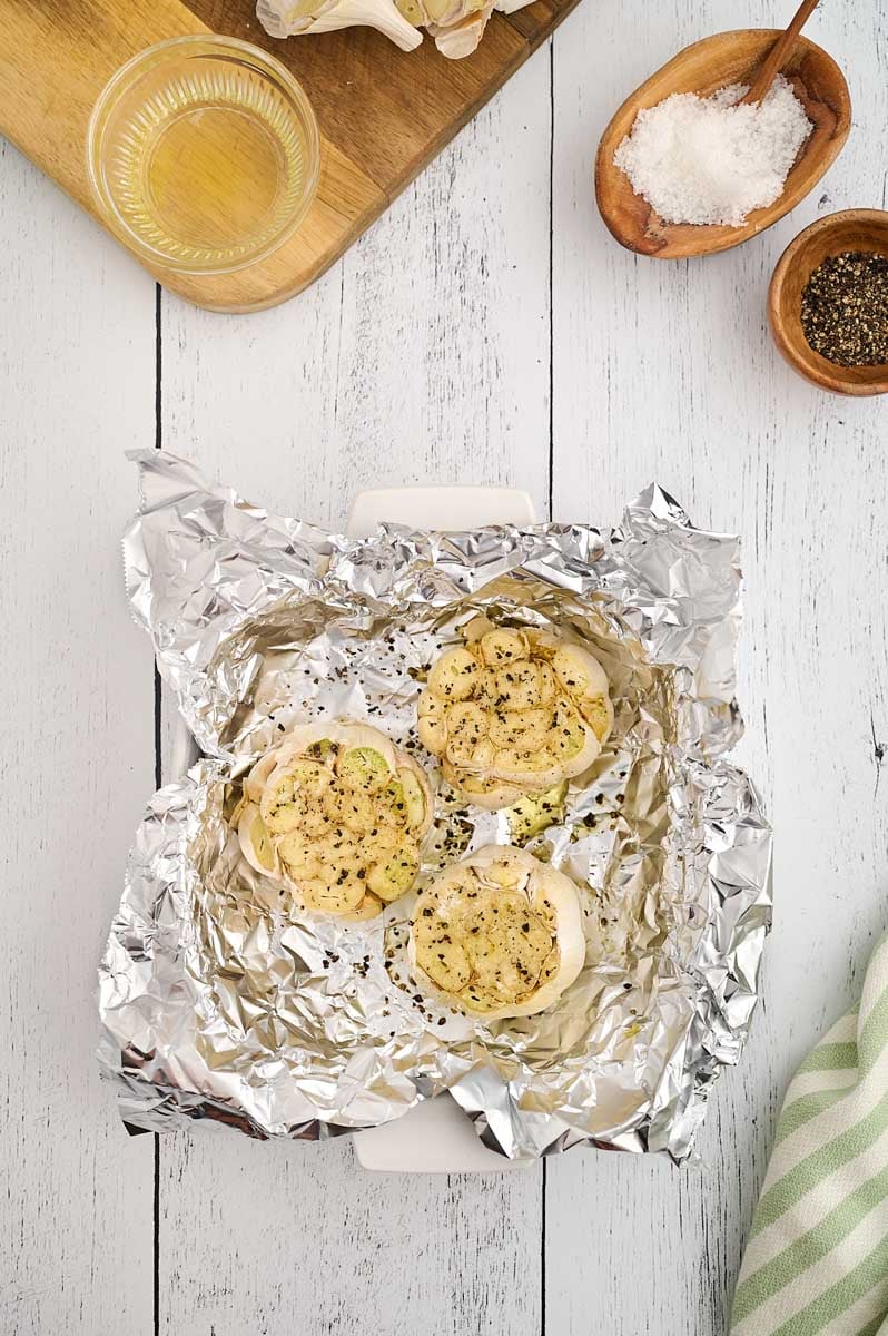 Roasted garlic bulbs covered in seasoning sit on a sheet of aluminum foil placed on a white wooden surface, with bowls of salt, pepper, and olive oil nearby.