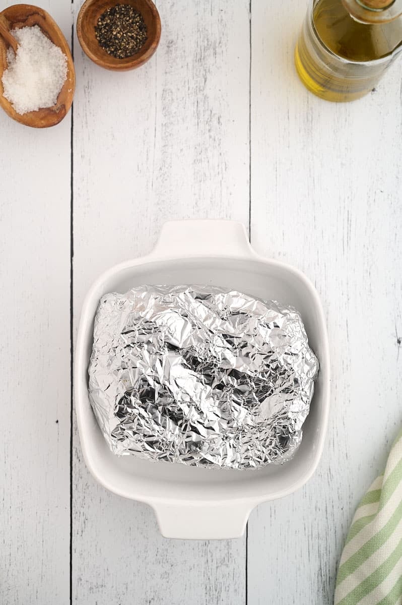 A foil-wrapped item sits in a white casserole dish on a white wooden surface. Nearby, there are bowls of salt and pepper, a bottle of olive oil, and a green and white striped cloth.