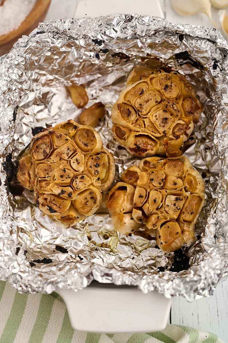 Three roasted garlic bulbs seasoned with black pepper in an aluminum foil-lined dish.