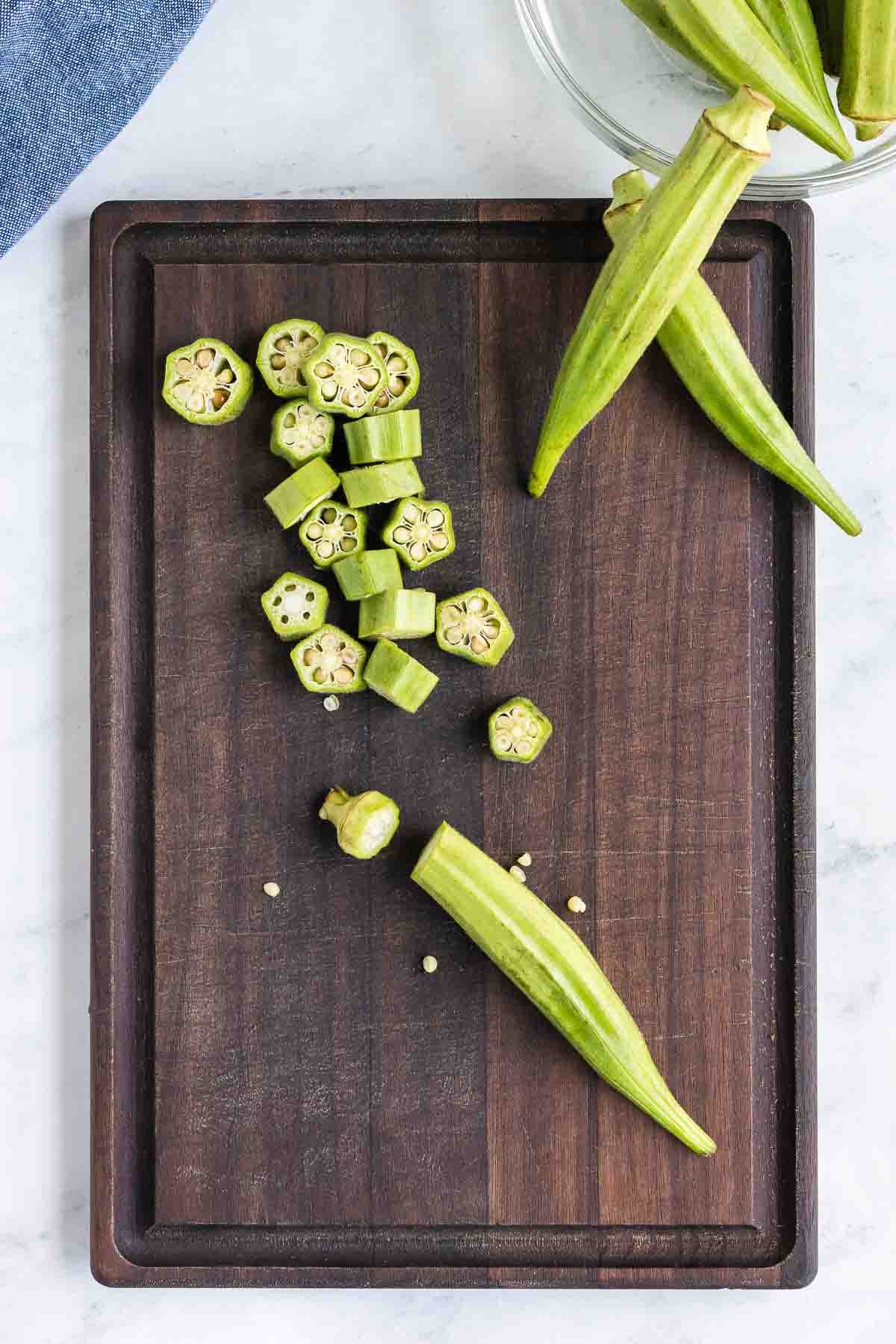Sliced and whole okra on a dark wooden cutting board with a blue cloth in the background. Some okra pieces have seeds visible.