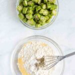 Two bowls on a marble surface: one with sliced okra and another containing a flour mixture with pepper being whisked in.