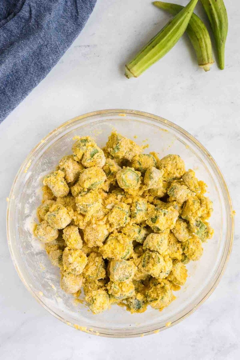 A clear glass bowl filled with seasoned, breaded okra pieces on a white countertop, with two whole okras and a dark blue cloth nearby.