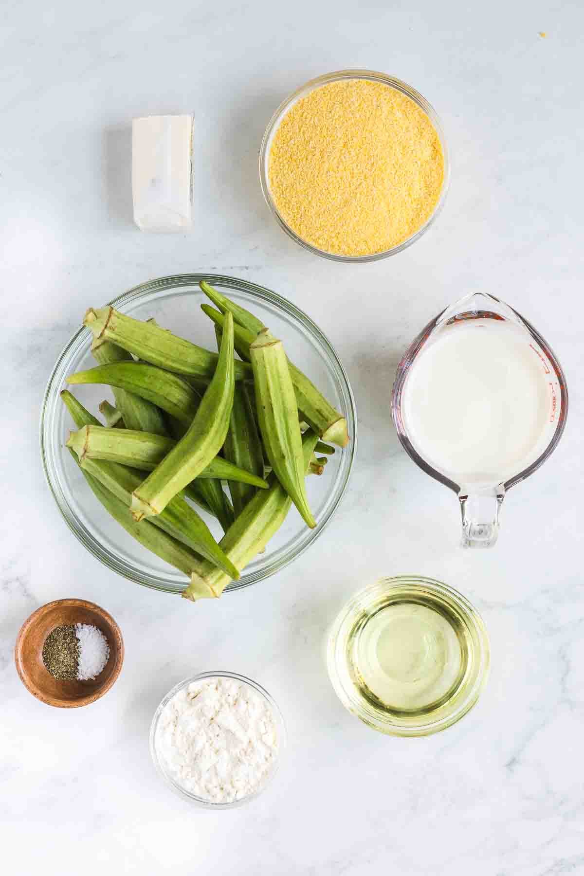 Top-down view of ingredients on a white surface including okra, milk, cornmeal, flour, cooking oil, butter, salt, and pepper.