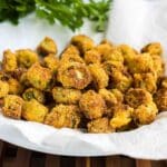 A plate of golden-brown fried okra sits on a white paper towel with fresh green herbs in the background.