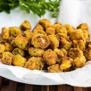 A plate of golden-brown fried okra sits on a white paper towel with fresh green herbs in the background.