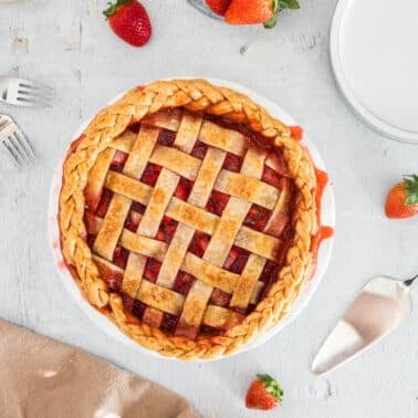 Top view of a freshly baked strawberry pie with lattice crust, surrounded by strawberries, forks, a pie server, and a folded brown cloth on a light surface.