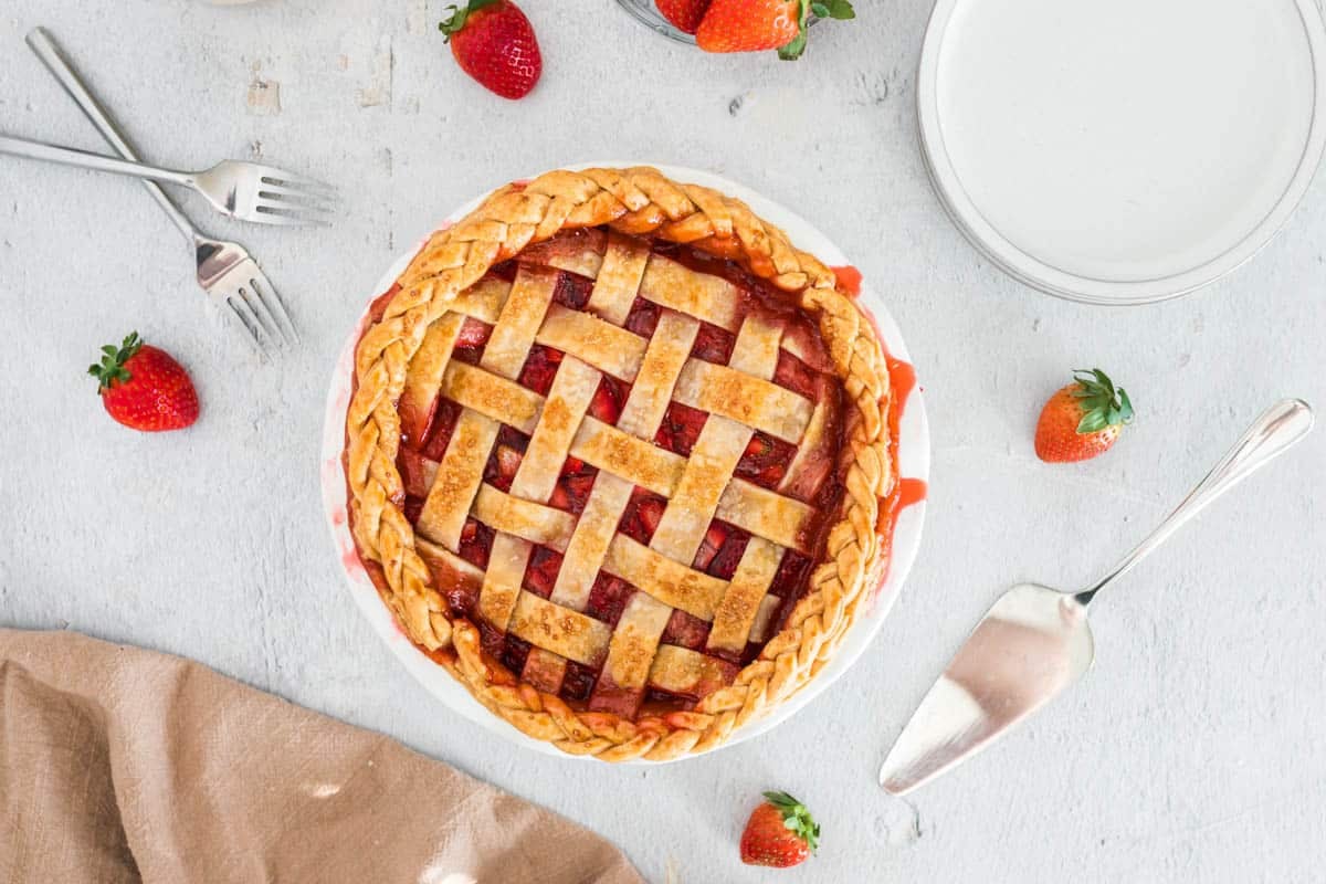 Top view of a freshly baked strawberry pie with lattice crust, surrounded by strawberries, forks, a pie server, and a folded brown cloth on a light surface.
