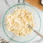 A glass bowl containing biscuit dough, with a fork partially mixing the contents. Nearby, there is a wooden board with a small container, a cloth with cursive print, and a bottle of milk.