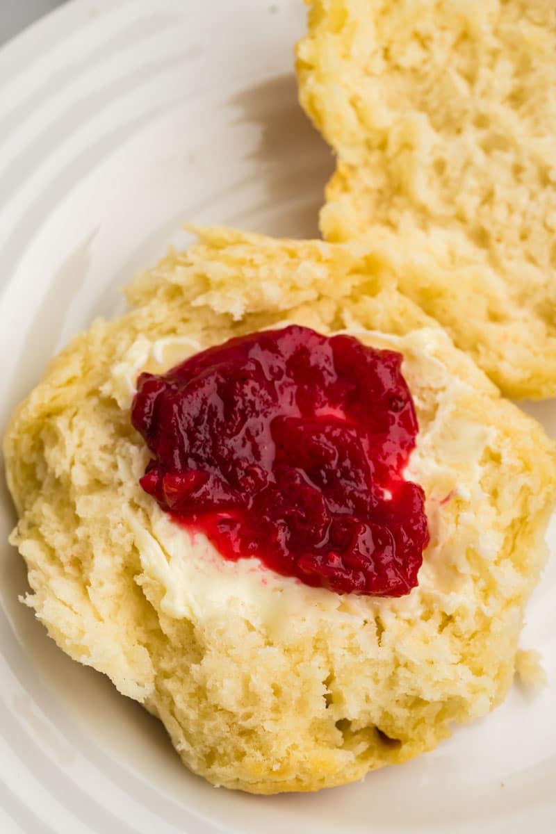 A biscuit split open with butter and strawberry jam on a white plate.