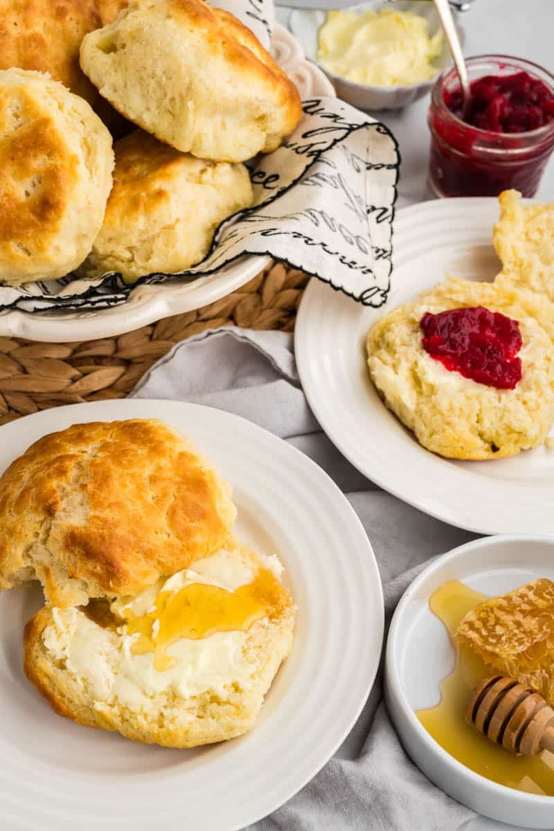 A basket of buttermilk biscuits on a woven mat, with two plates each featuring a biscuit; one with honey and butter, and the other with butter and red jam. Honeycomb and butter in background.