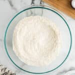 A glass bowl filled with flour is on a marble surface. Nearby are a dish with butter cubes, a milk bottle, a small bowl with salt, and a wooden board. A patterned cloth is partially visible.