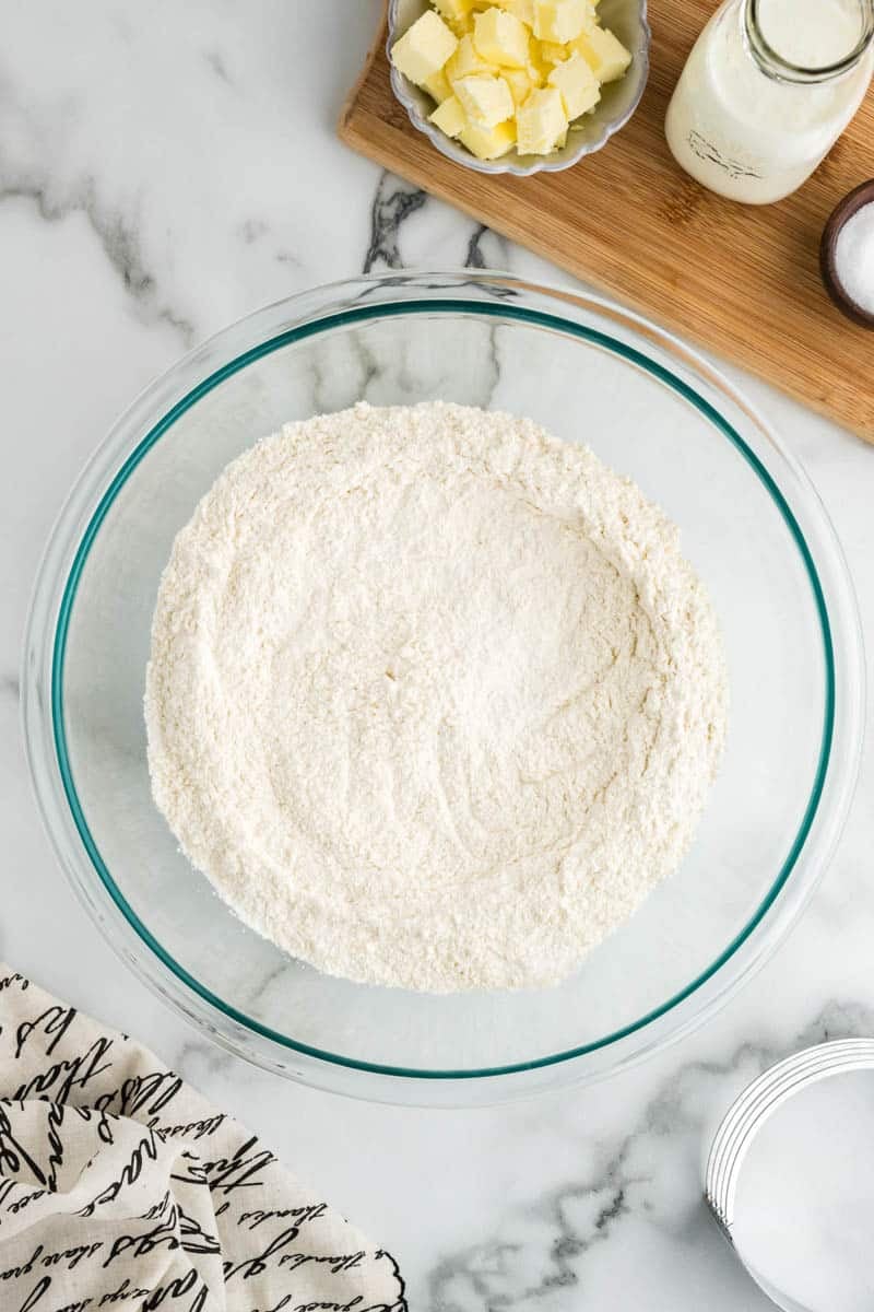 A glass bowl filled with flour is on a marble surface. Nearby are a dish with butter cubes, a milk bottle, a small bowl with salt, and a wooden board. A patterned cloth is partially visible.