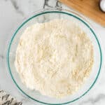 A large glass bowl filled with flour and butter mixture on a marble countertop, next to a wooden board with a small bottle of cream and a pinch bowl of salt. A cloth with text is partially visible.
