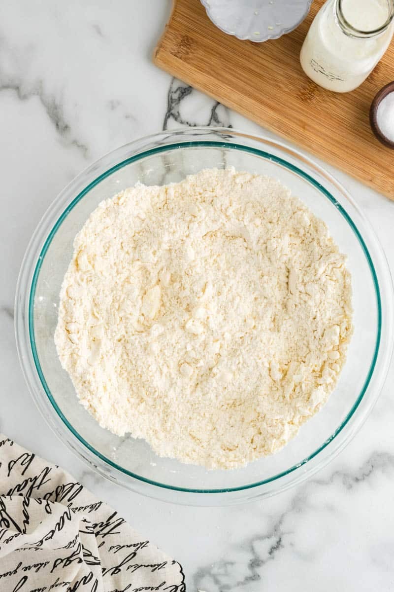 A large glass bowl filled with flour and butter mixture on a marble countertop, next to a wooden board with a small bottle of cream and a pinch bowl of salt. A cloth with text is partially visible.