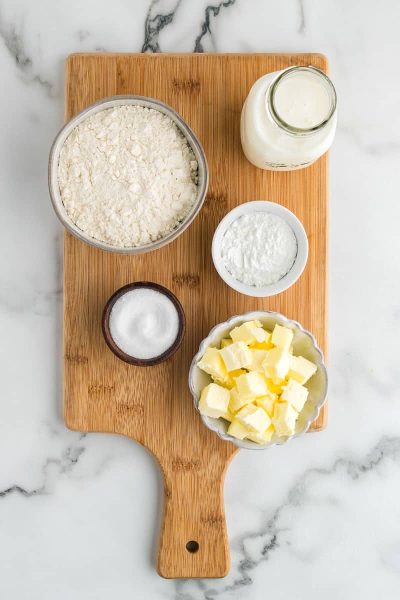 A wooden cutting board with a bowl of flour, a small bowl of sugar, a bowl of cubed butter, a container of milk, and a small dish of baking powder. All ingredients are set on a marble surface.