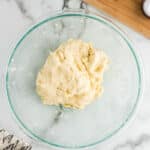 A glass bowl with dough placed on a marble countertop. A wooden cutting board, small white bowl, flour canister, and hand towel are seen in the background.