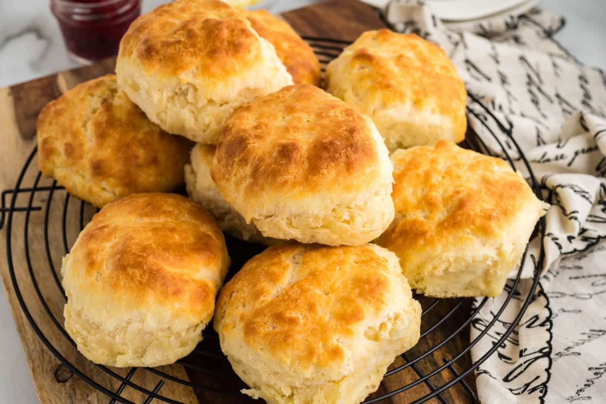 A group of golden-brown buttermilk biscuits is arranged on a round cooling rack, placed on a wooden cutting board. A patterned napkin is partially visible on the right side.