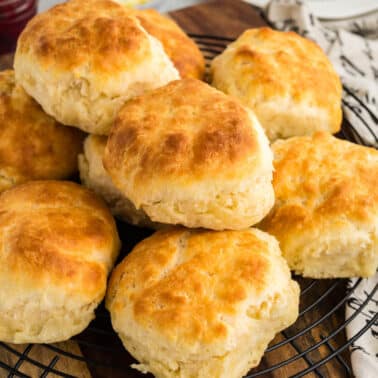 A stack of freshly baked golden-brown biscuits on a cooling rack, placed on a wooden board next to a cloth with writing and a jar of jam.