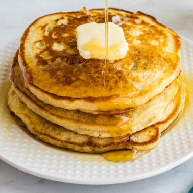 A stack of pancakes topped with butter and syrup on a white plate, accompanied by a glass of milk in the background.