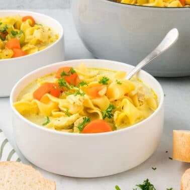 Bowl of chicken noodle soup with carrots and parsley, served with bread slices. A larger pot and another bowl of soup are in the background, next to salt and pepper shakers.