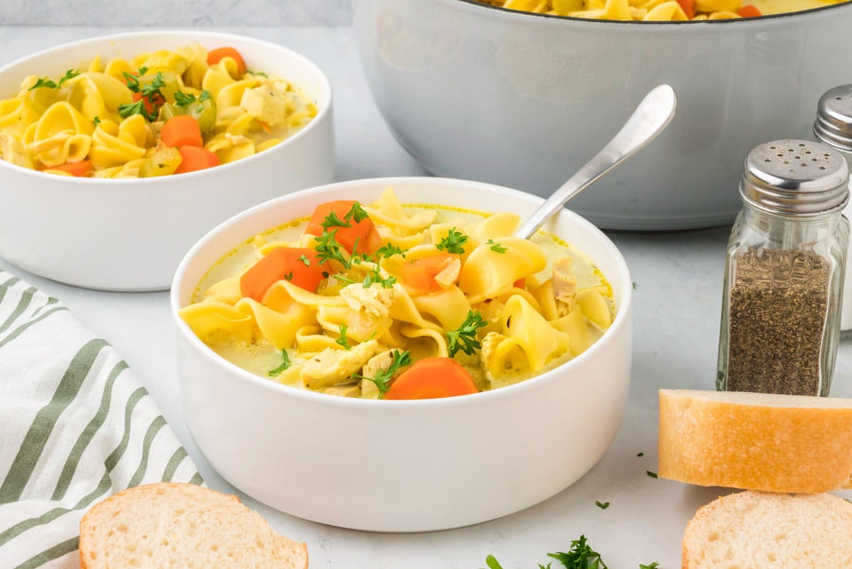 Bowl of chicken noodle soup with carrots and parsley, served with bread slices. A larger pot and another bowl of soup are in the background, next to salt and pepper shakers.