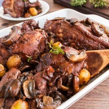 A white dish filled with Julia Child's Coq au Vin, on a wooden table. A wooden spoon rests on the edge of the dish.