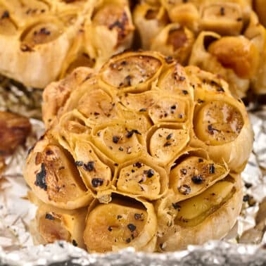 Close-up of roasted garlic bulbs with cloves visible, seasoned with black pepper and herbs, resting on aluminum foil.