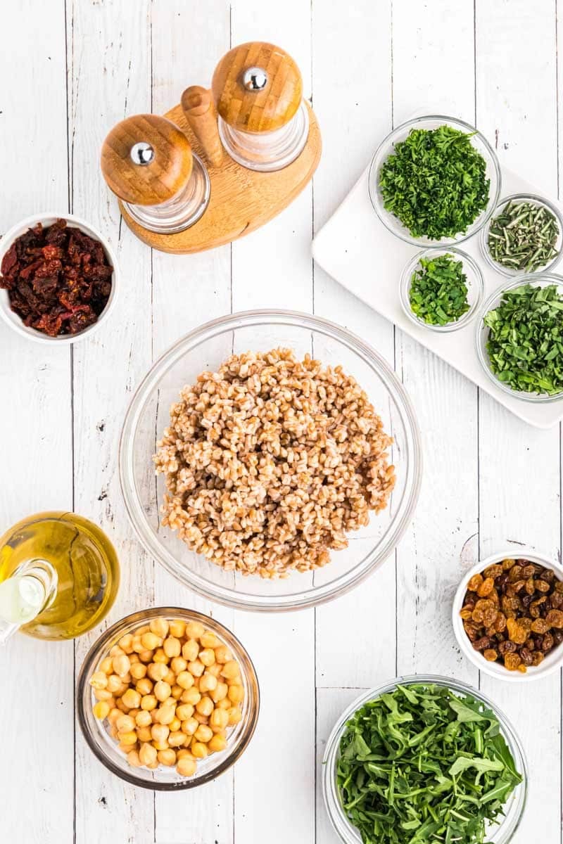 A top-down view of ingredients for a salad, including cooked grains, chickpeas, dried fruit, sun-dried tomatoes, herbs, arugula, olive oil, and salt and pepper shakers on a white wooden surface.