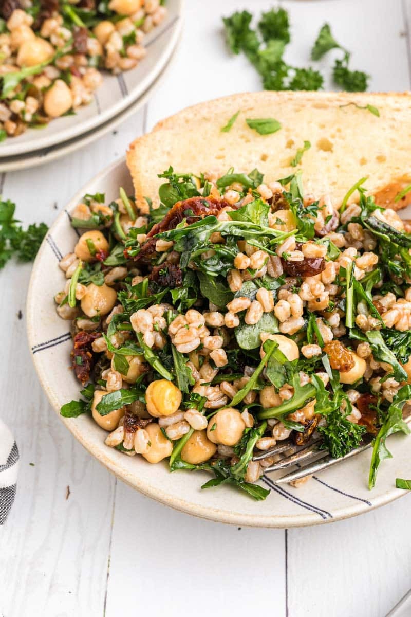 A bowl of mixed salad containing chickpeas, green leafy vegetables, farro, and sun-dried tomatoes, accompanied by a slice of bread.