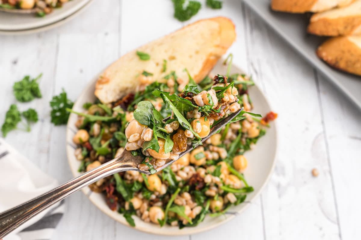 A fork holds a portion of a farro arugula salad over a white plate, garnished with a slice of toasted bread. In the background, more bread slices and a partial salad are visible.