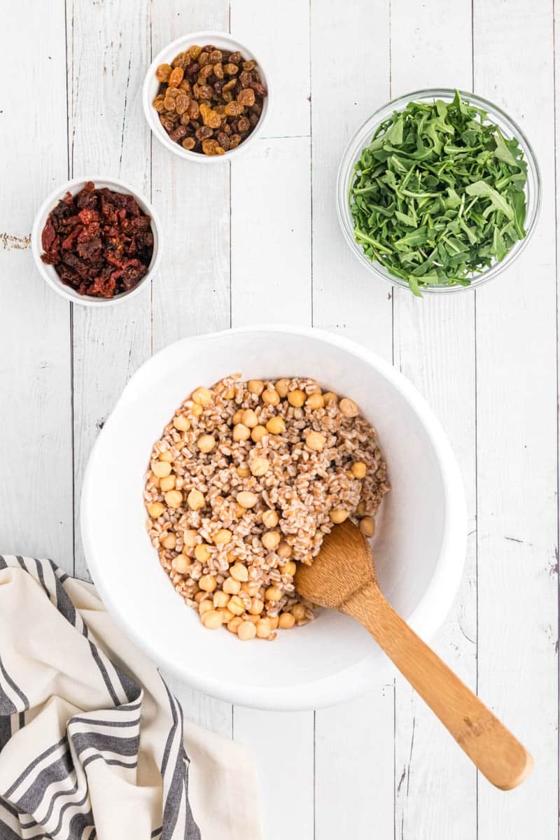 A white bowl with a mixture of chickpeas and grains, next to three smaller bowls containing arugula, dried tomatoes, and raisins on a white wooden surface, with a striped cloth nearby.