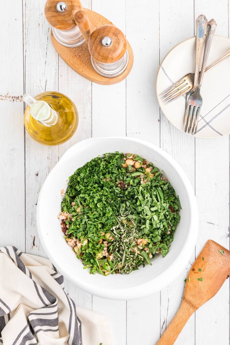 A white bowl filled with greens and a mix of ingredients on a white wooden table, accompanied by a wooden spatula, a striped cloth, olive oil bottle, and plates with forks.