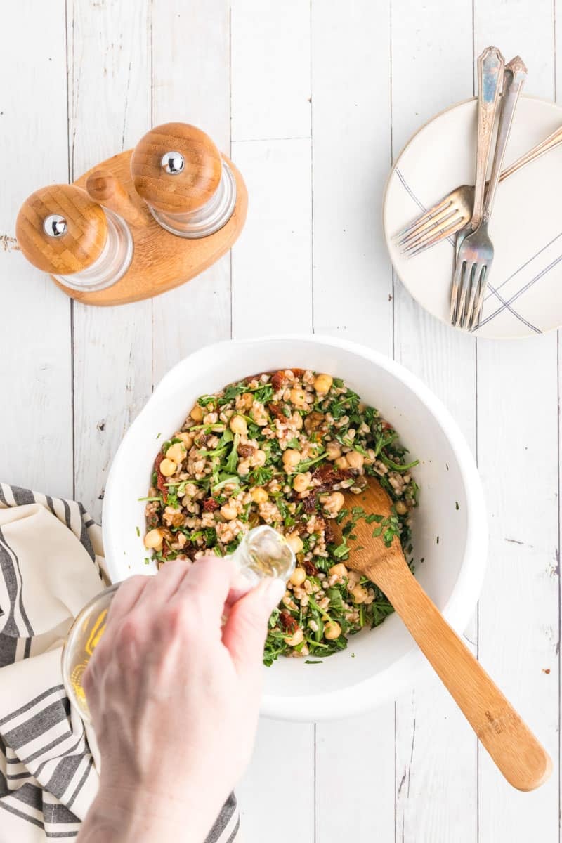 A hand pouring oil into a bowl of mixed salad with greens and beans, surrounded by wooden salt and pepper shakers, plates with forks, and a striped cloth on a white wooden surface.