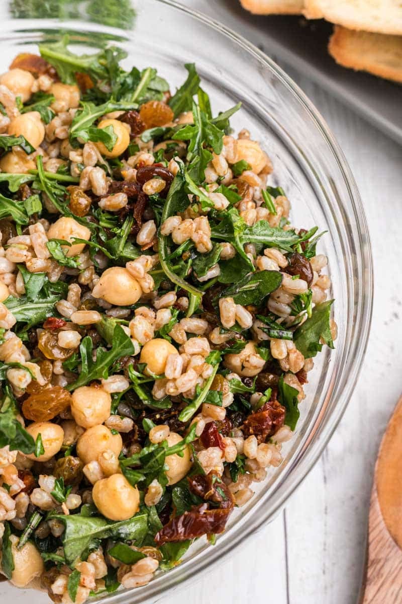 A clear glass bowl containing a mixed salad of chickpeas, farro, arugula, raisins, and herbs. A plate of sliced bread is partially visible in the background.