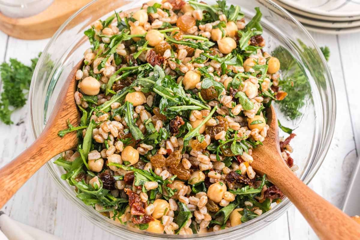 A mixed salad in a glass bowl containing chickpeas, farro, sun-dried tomatoes, arugula, and golden raisins is being served with wooden salad tongs.