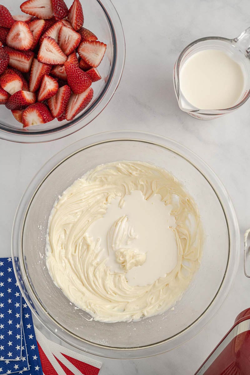 A mixing bowl with partially mixed cream cheese and cream, beside a bowl of sliced strawberries, a container of cream, and an American-themed napkin.