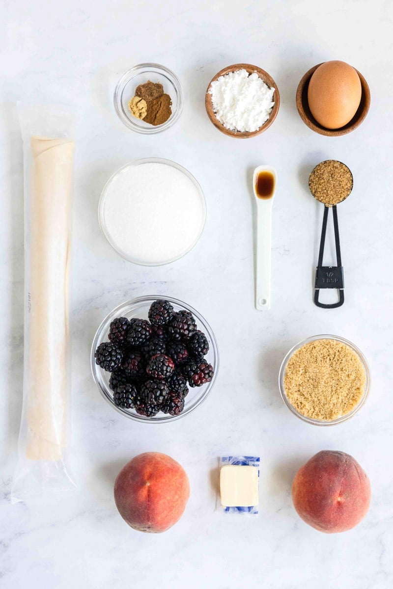 Ingredients laid out on a countertop, including blackberries, peaches, a sheet of dough, sugar, spices, an egg, vanilla extract, cornstarch, butter, and measuring spoons.