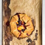 A rustic fruit galette with a golden brown crust sits on a crumpled piece of parchment paper on a baking sheet. Some fruit filling has oozed out and caramelized on the paper and baking sheet.