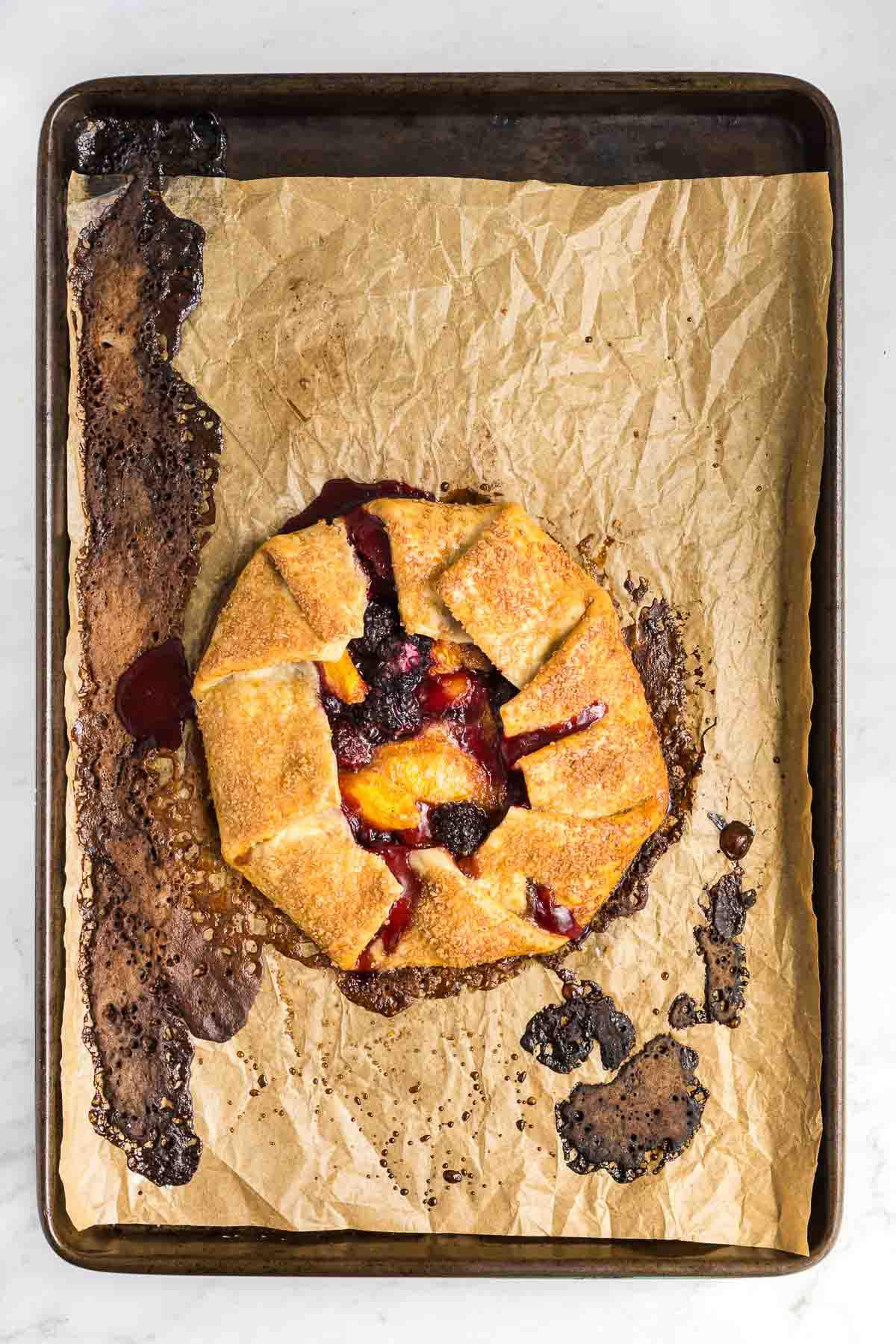 A rustic fruit galette with a golden brown crust sits on a crumpled piece of parchment paper on a baking sheet. Some fruit filling has oozed out and caramelized on the paper and baking sheet.