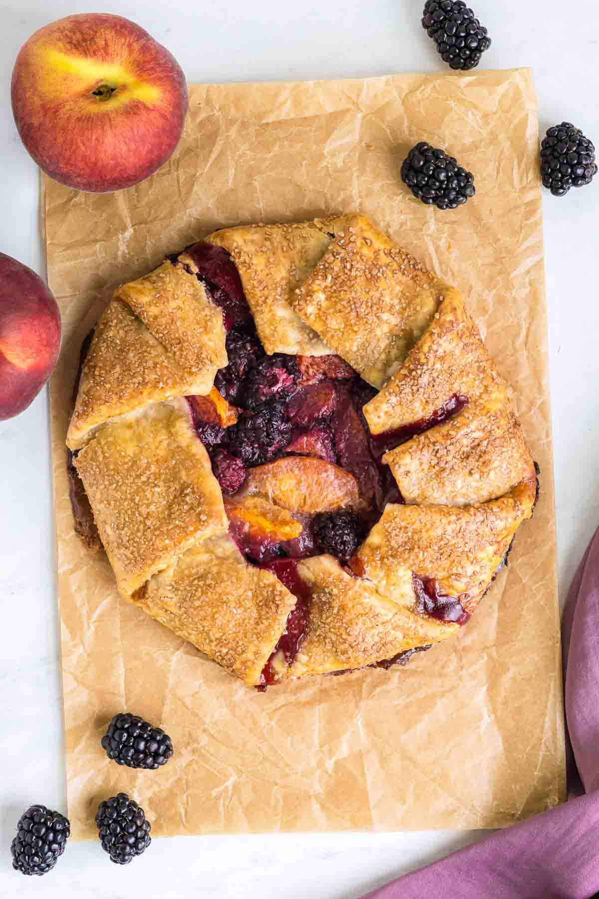 A rustic peach and blackberry galette on parchment paper, surrounded by fresh blackberries and peaches.