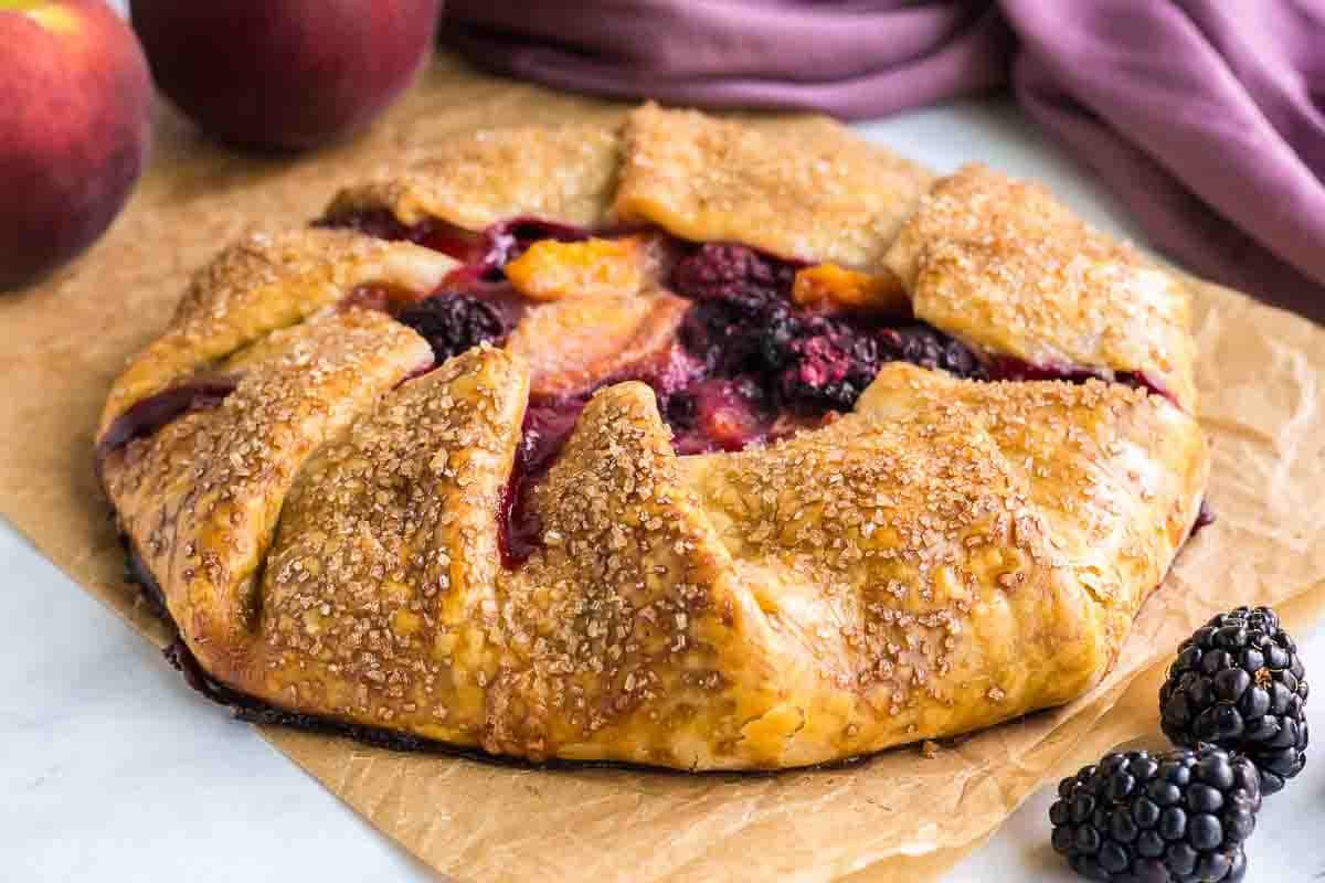 A rustic peach blackberry galette with a golden, flaky crust filled with mixed berries and peach slices, on a piece of parchment paper. A few fresh blackberries and peaches are placed nearby.