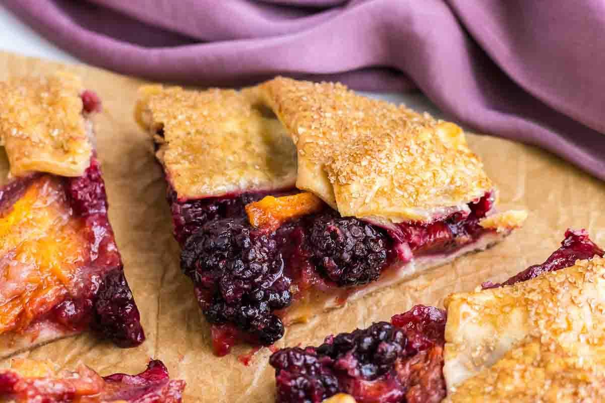 Close-up of slices of peach blackberry galette with a golden, flaky crust, placed on a parchment paper, with a purple cloth in the background.